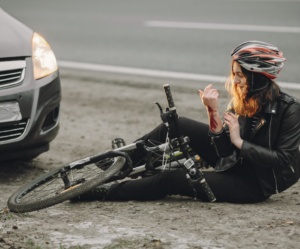 A cyclist wearing a helmet and a black jacket sits injured on the ground next to a car, holding her bloody arm